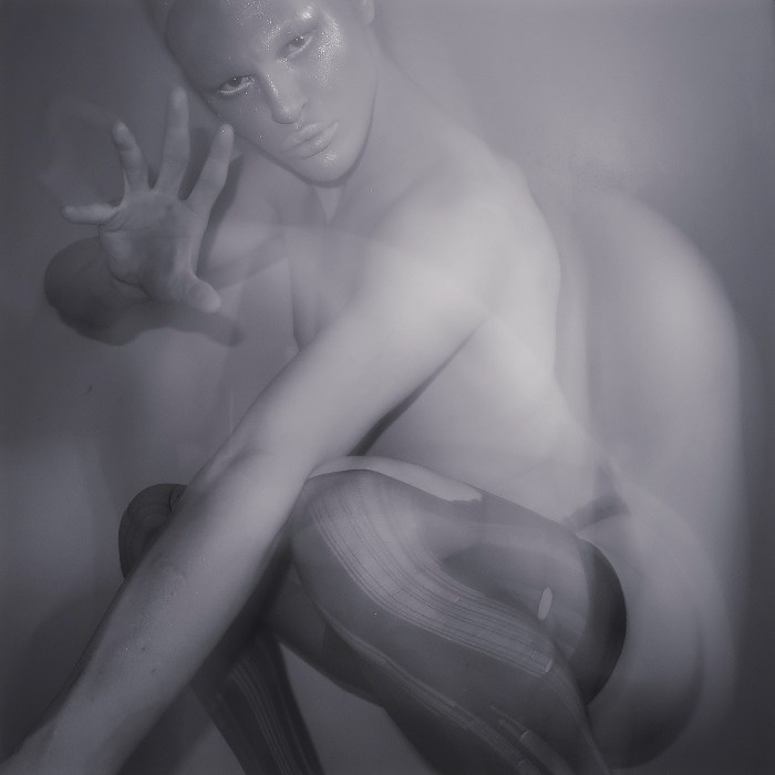 A black and white photo of a crouching person wearing shimmering make-up. They are wearing ripped stockings and reaching toward the camera. 