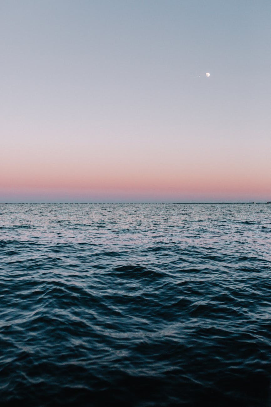 Image Description: A body of water with small waves at dusk. The horizon fades from powdery blue to powdery pink. A gibbous moon glows in the right corner of the sky.