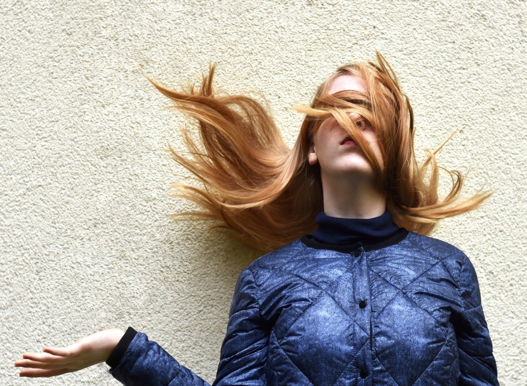 A woman stands against a white wall with her long hair blowing up around her face and covering her eyes. Her right arm is bent at the elbow with the palm upturned, as if waiting for something or asking a question.