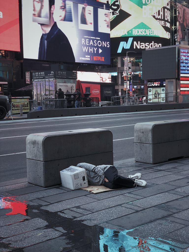 City street lined with billboards. A person lies on the sidewalk in the fetal position on a piece of cardboard with a box over their head.
