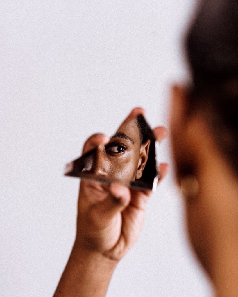 A black person (gender unknown) holds up a sliver of mirror that reflects a portion of their face.