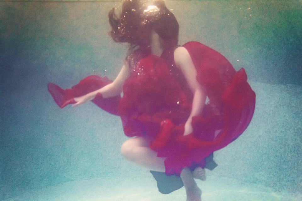 A woman in a red dress floats underwater. Her long hair covers her face as it rises towards the surface. 