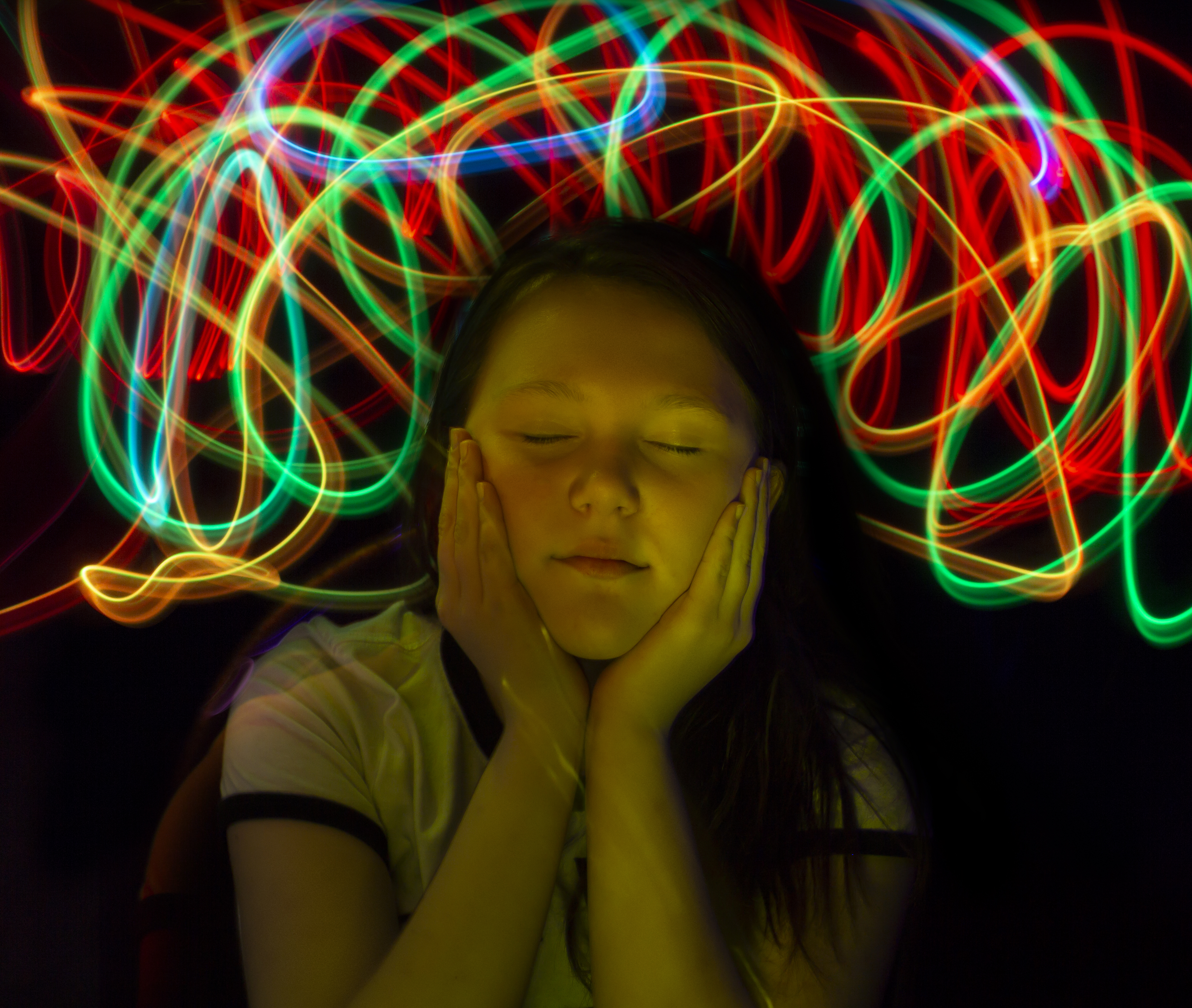 A girl with her eyes closed cups her cheeks with her hands in the dark under neon swirls.